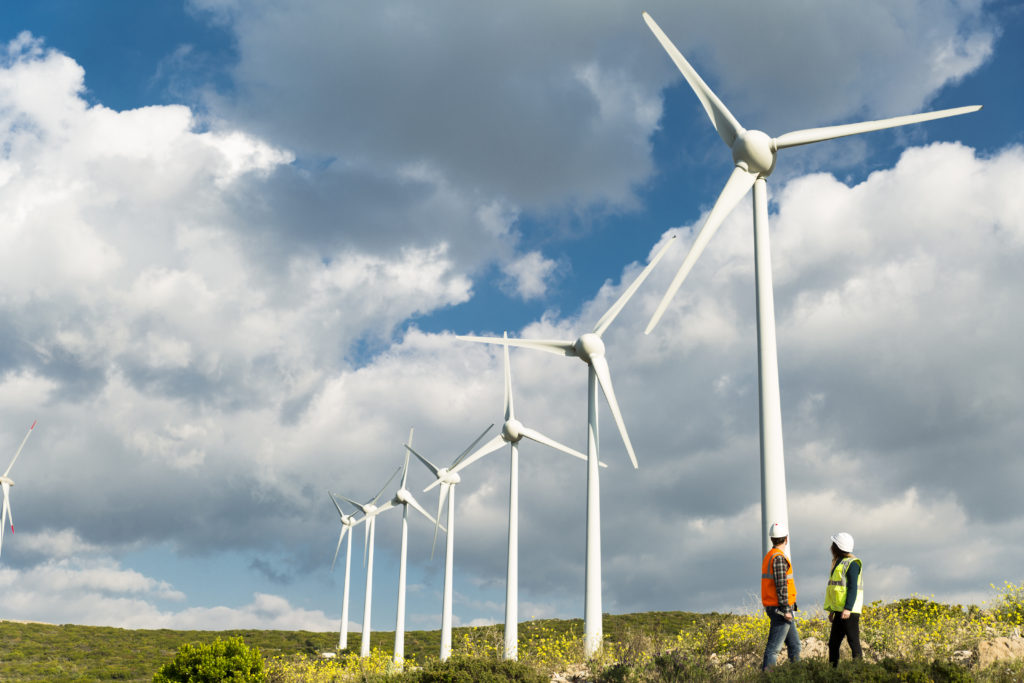 Two young caucasian technician engineer man and woman wearing white worker headwear standing, working, checking farm field system and looking up at wind turbine farm power generation station in rural landscape.